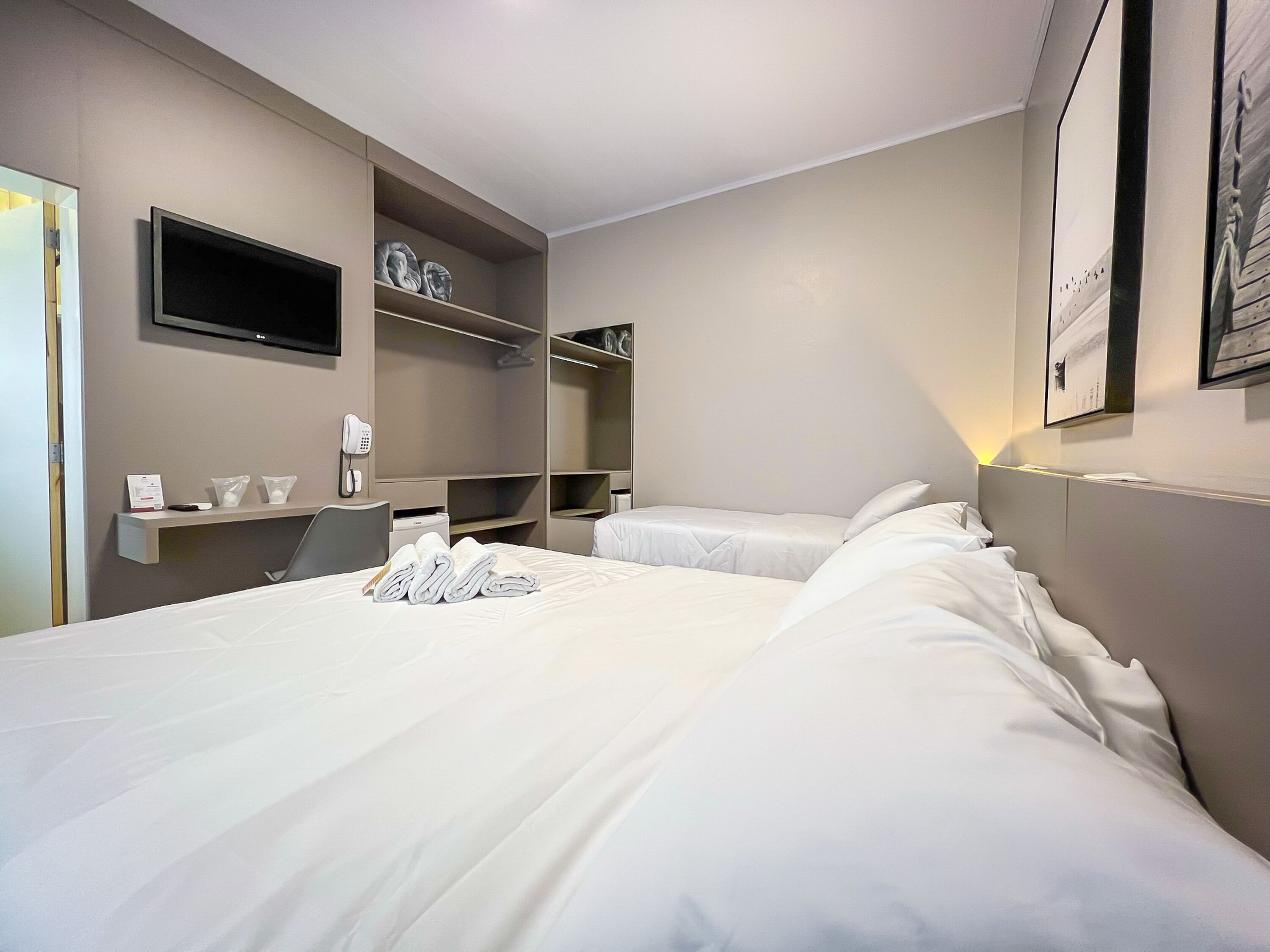 A hotel room with a large bed, a desk with a chair, a telephone, and a flat-screen TV mounted on the wall.