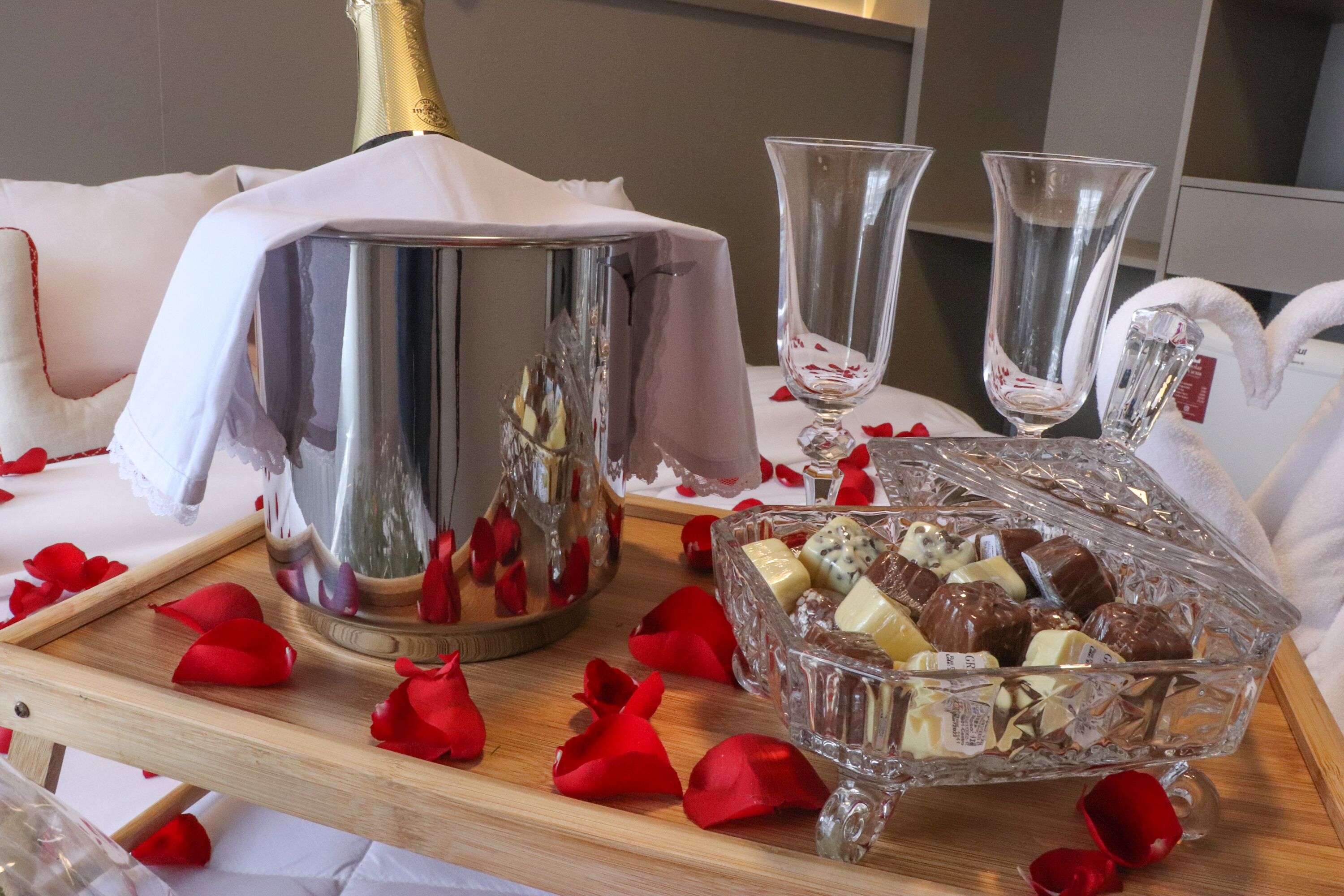 A table with a champagne bottle, two glasses, and a glass bowl of chocolates, surrounded by rose petals.