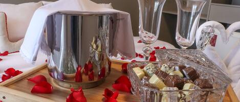A table with a champagne bottle, two glasses, and a glass bowl of chocolates, surrounded by rose petals.