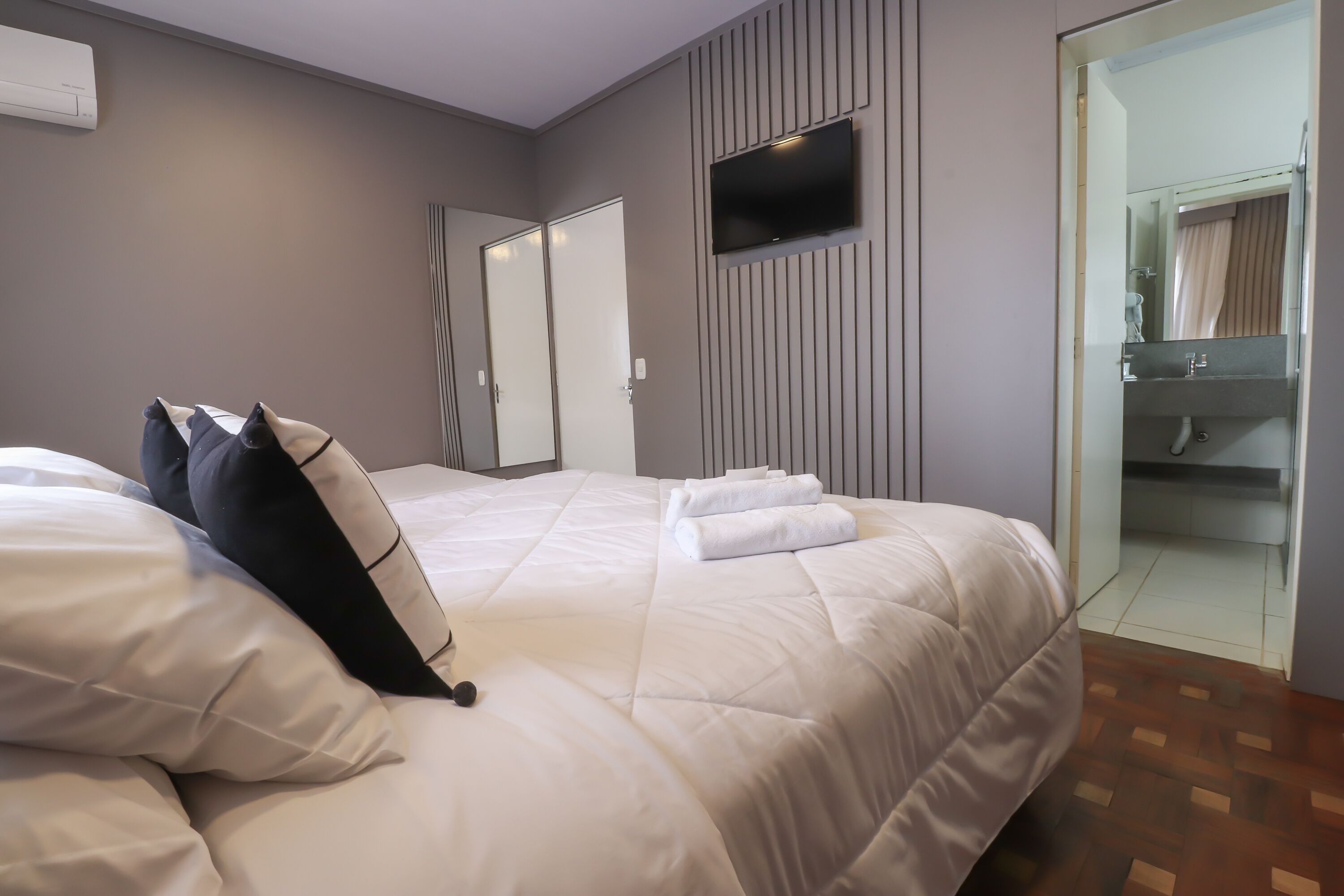 A neatly made bed with white and black pillows, a flat-screen TV mounted on the wall, and a bathroom visible through a glass door.