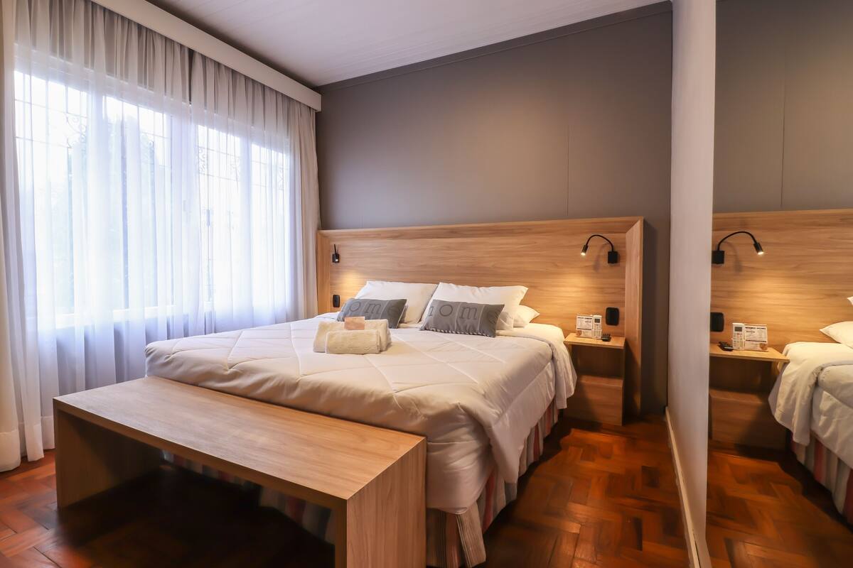 Une chambre d’hôtel moderne avec un grand lit, des tables de chevet, un banc et une vue sur l’extérieur à travers des rideaux transparents.