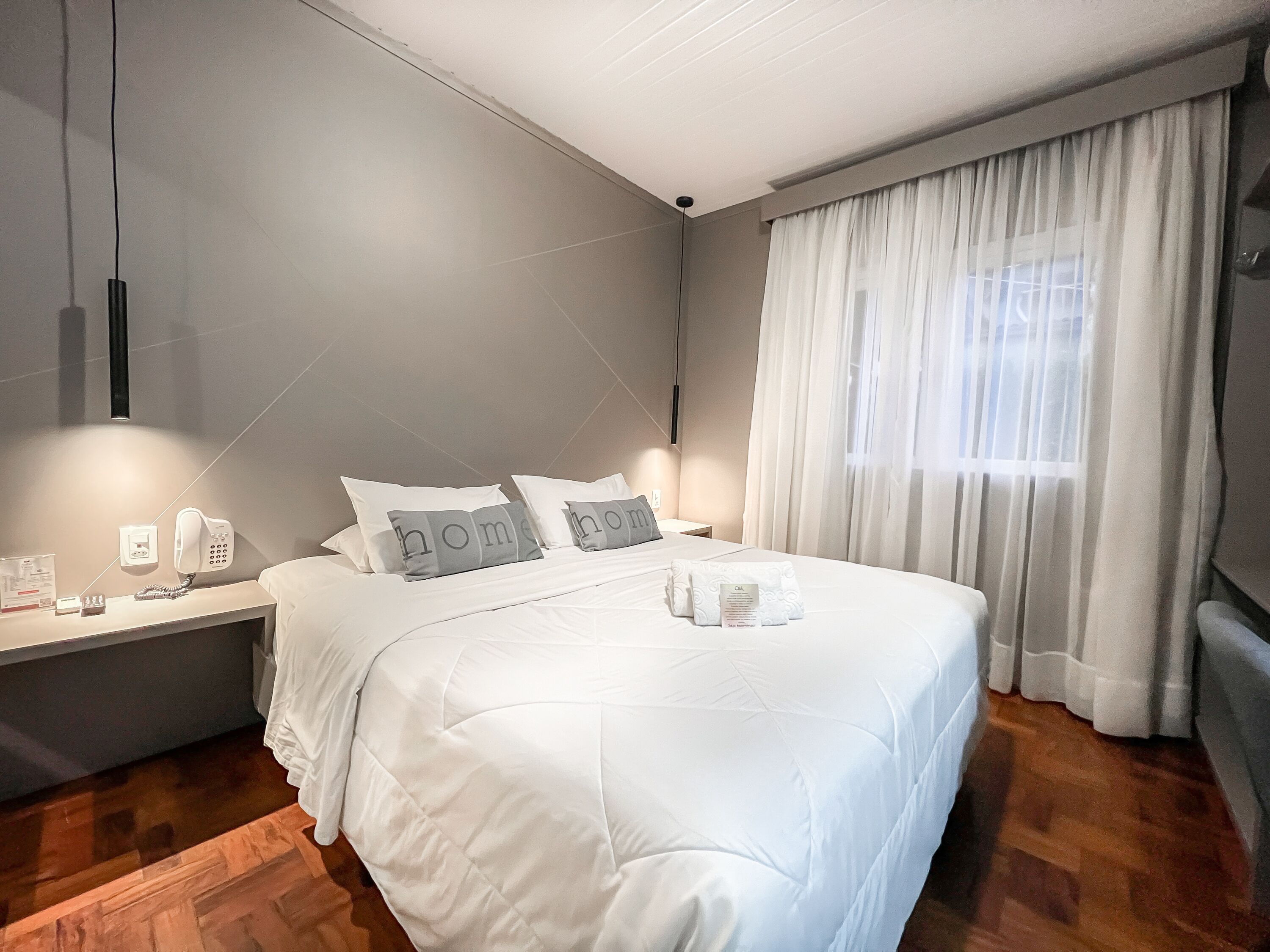 A neatly made bed with white bedding and pillows, a bedside table with a phone and a lamp, and a window with sheer curtains.