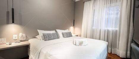 A neatly made bed with white bedding and pillows, a bedside table with a phone and a lamp, and a window with sheer curtains.