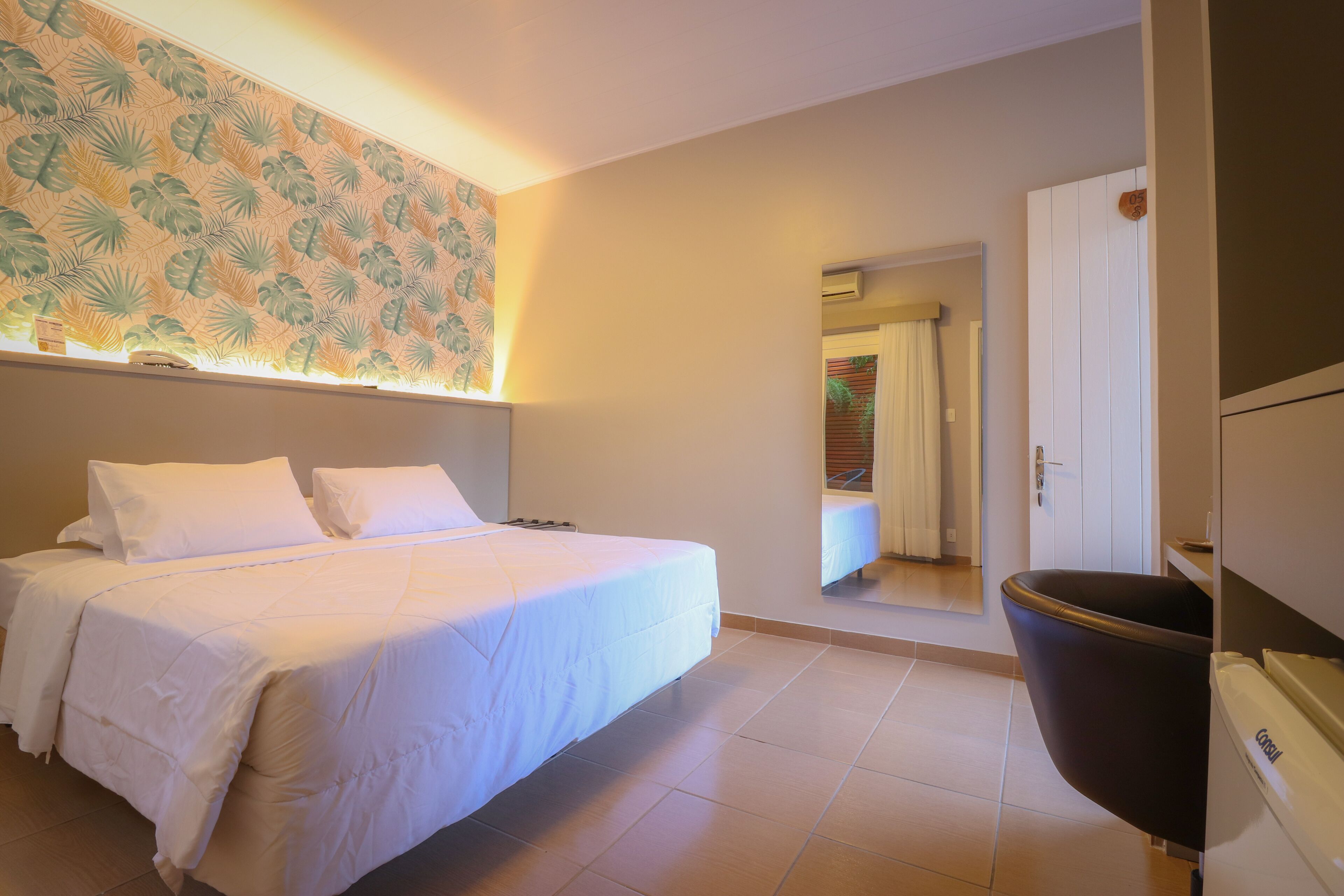 A bedroom with a bed, a nightstand, a wall-mounted lamp, and a bathroom door visible in the background.