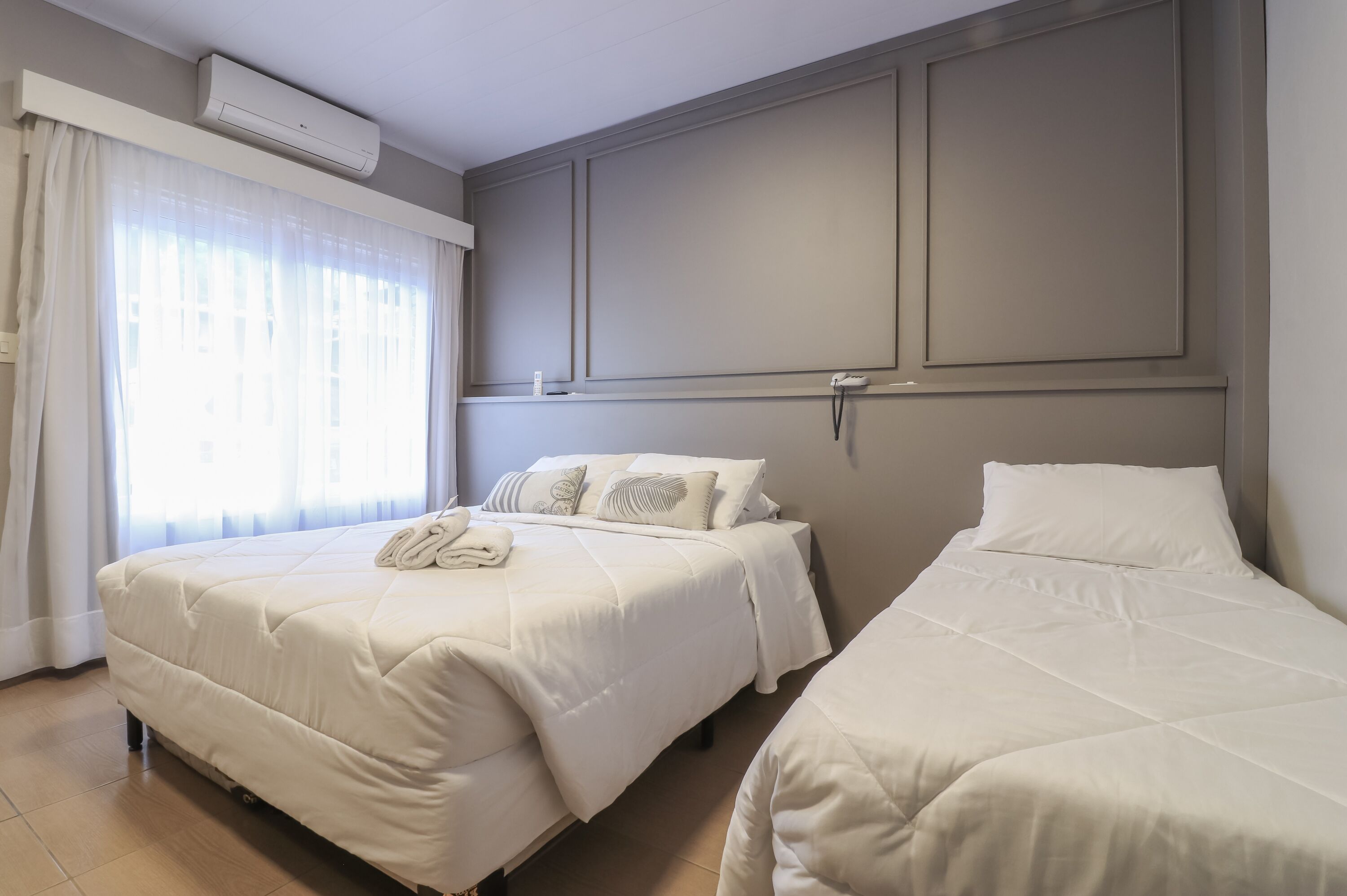 A hotel room with two beds, a large window with sheer curtains, a wall-mounted air conditioner, and a plain, neutral-colored wall.