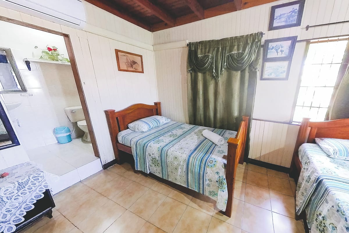 Una habitación pequeña y ordenada con dos camas individuales, un cabecero de madera, una mesita con mantel de encaje y una ventana con cortinas.