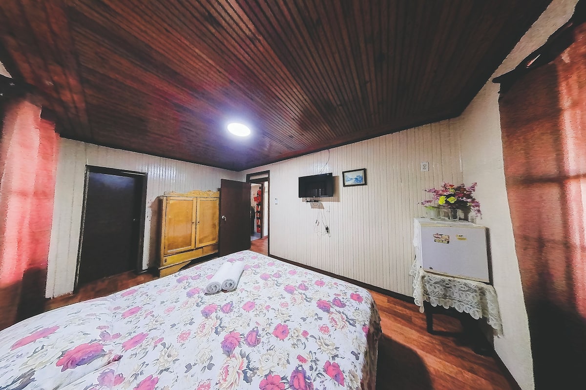Un dormitorio con techo de madera, una cama con ropa de cama floral, un armario, un televisor y una mesita de noche con un florero.