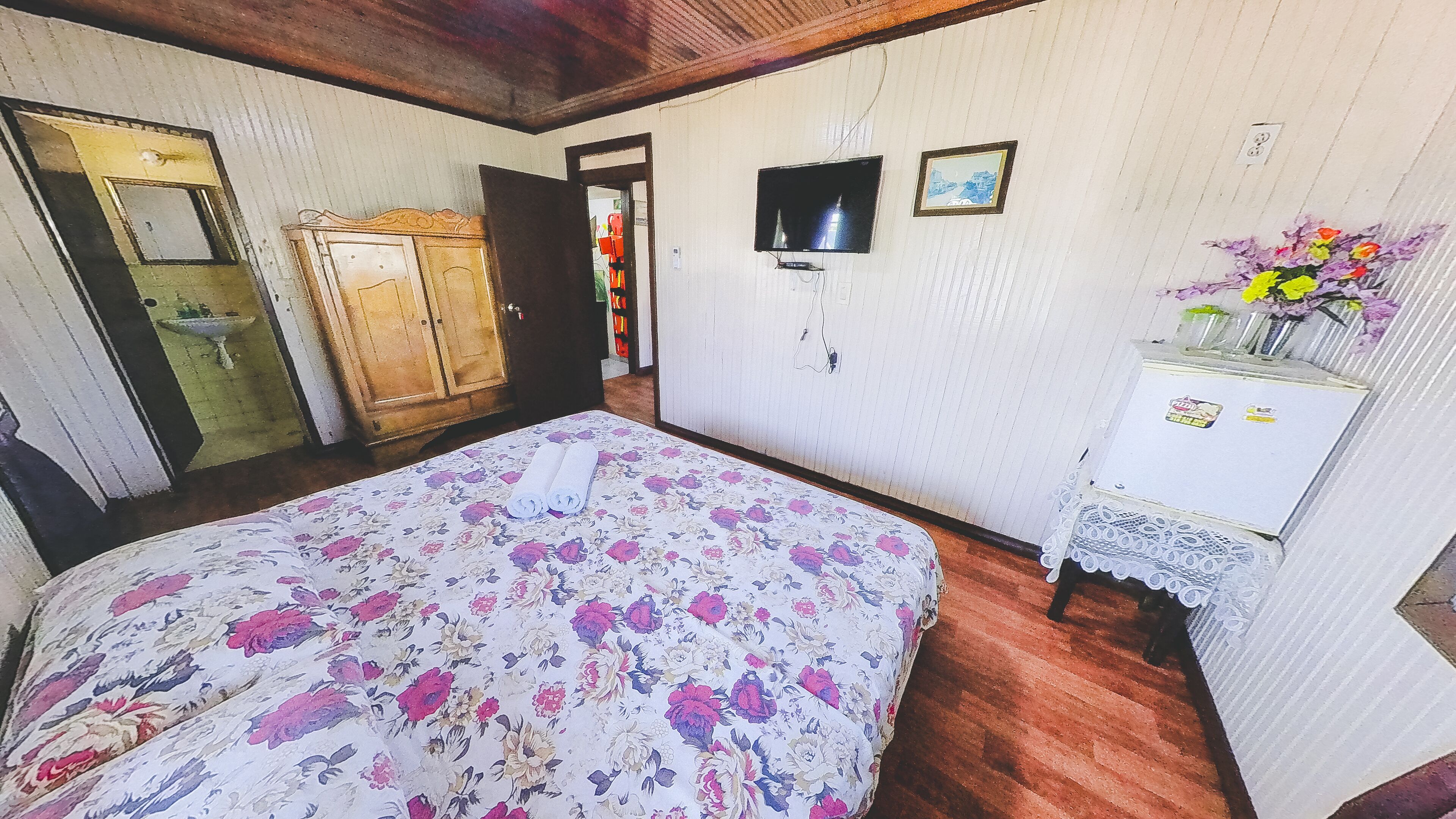 A bedroom with a floral bedspread, wooden wardrobe, and a small white cabinet with a vase of flowers.