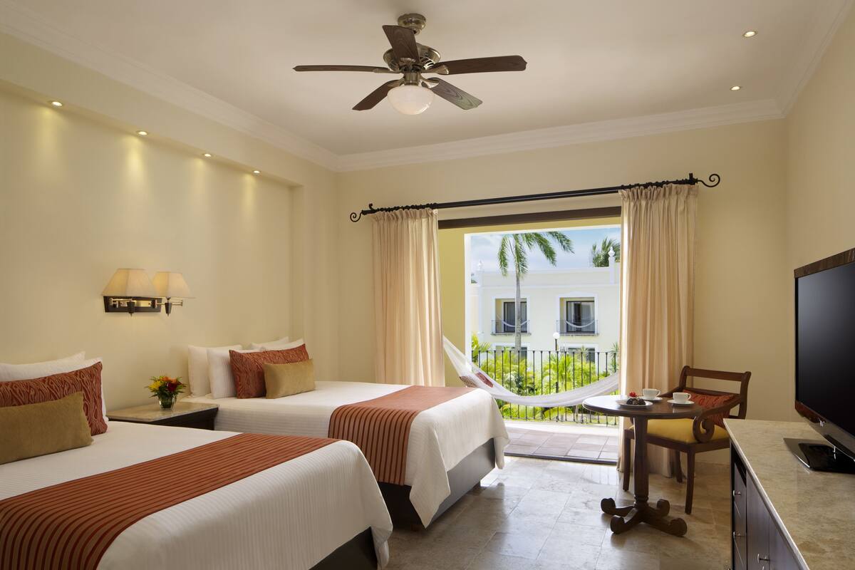 A hotel room with two beds, a television, a dining table, and a balcony with a view of palm trees.