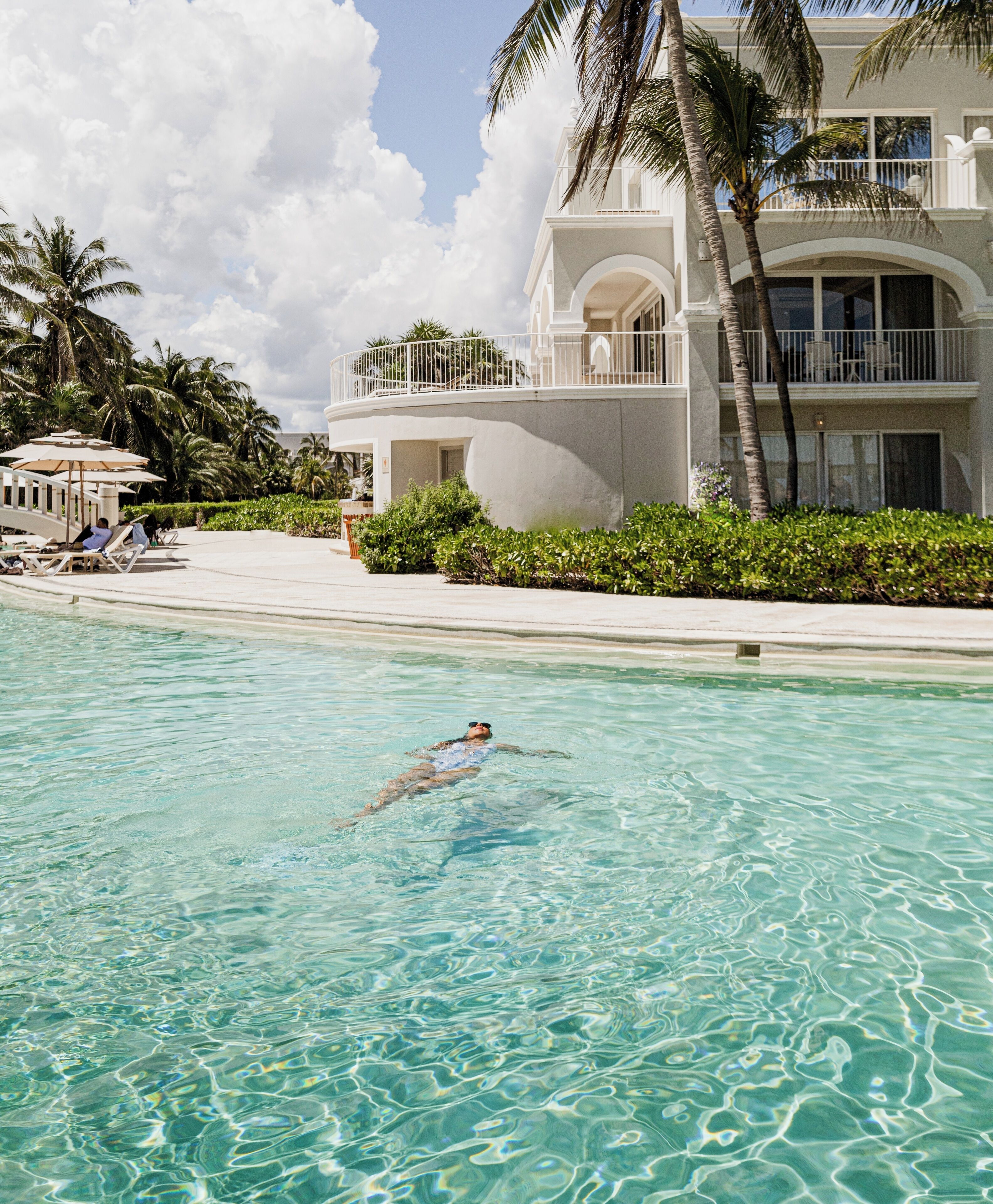 A swimming pool with clear water, a person swimming, and a resort building with balconies and palm trees.