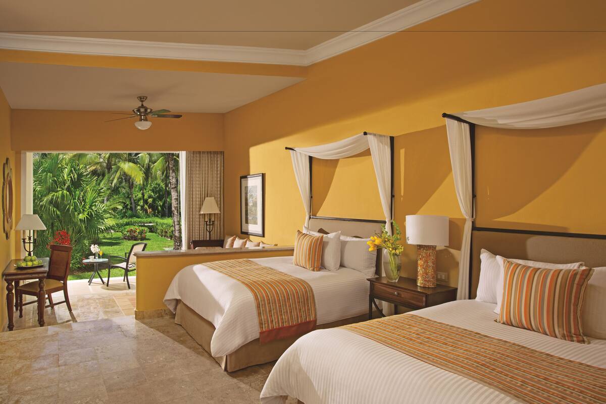 A hotel room with two beds, a sitting area with a table and chairs, and a view of a tropical garden.