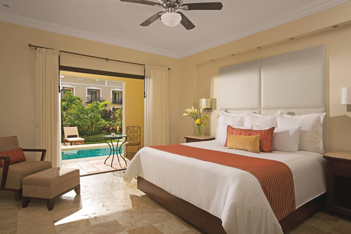 A hotel bedroom with a large bed, a ceiling fan, a view of a pool and greenery outside, and a chair by the window.