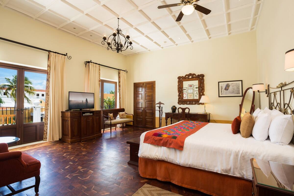 A hotel room with a large bed, a TV, a wooden desk, a chair, a ceiling fan, and a view of palm trees.