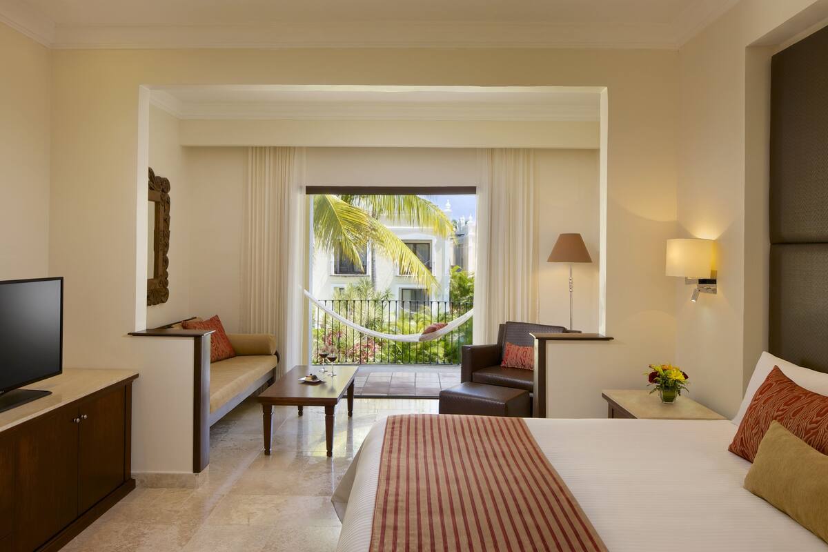 A hotel room with a bed, a desk, a chair, a television, and a view of a palm tree and building through a window.