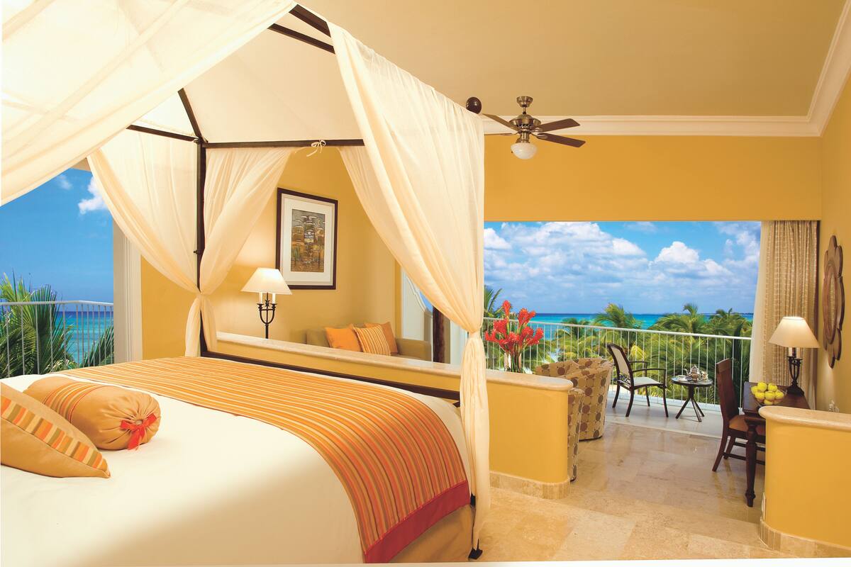 A bedroom with a four-poster bed, a balcony with a view of the ocean, and a ceiling fan.