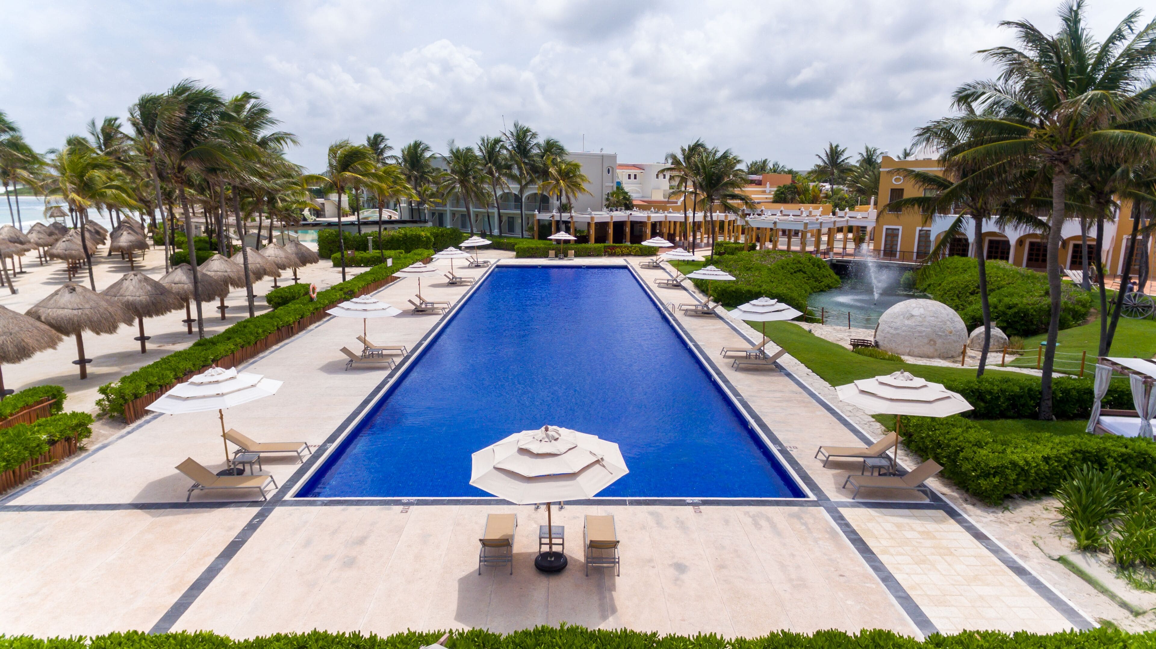 A large rectangular swimming pool with lounge chairs and umbrellas around it, surrounded by palm trees and a small waterfall.