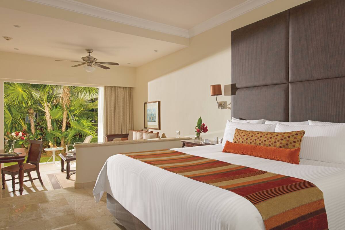 A spacious hotel room with a large bed, a balcony with a view of palm trees, a ceiling fan, and a small table with chairs.