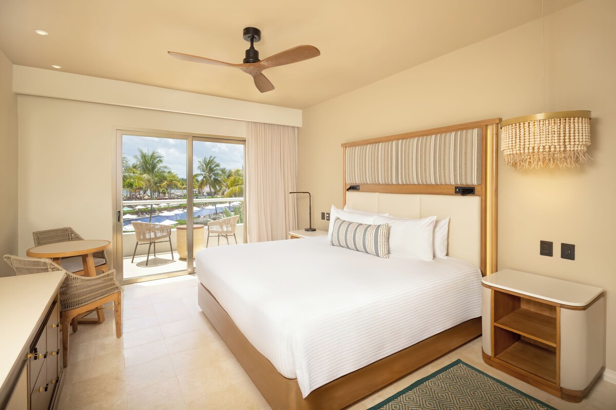 A hotel room with a bed, a desk, a chair, a ceiling fan, and a view of a pool and palm trees.