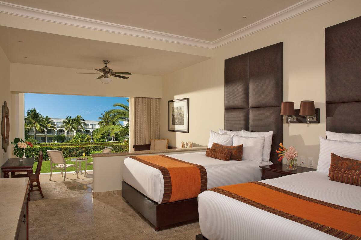 A hotel room with two beds, a balcony with a view of palm trees, a ceiling fan, and a desk with a chair.