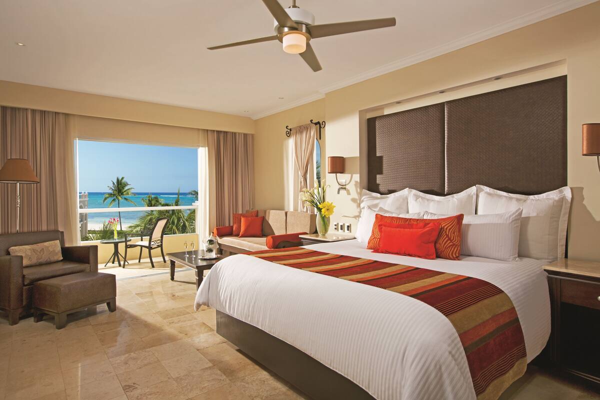 A hotel room with a large bed, a balcony with a view of the ocean, a seating area with a table, and a ceiling fan.