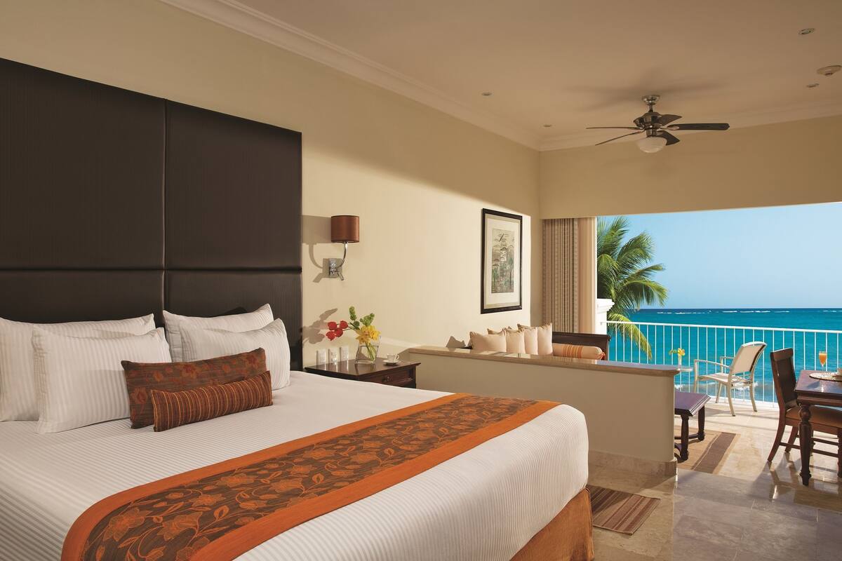 A hotel room with a large bed, a balcony with a view of the ocean, and a dining area with a table and chairs.