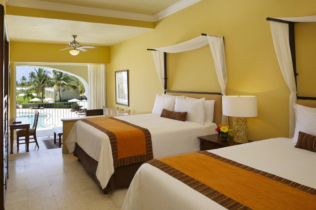 A hotel room with two beds, a balcony with a view of palm trees, and a dining area with a table and chairs.
