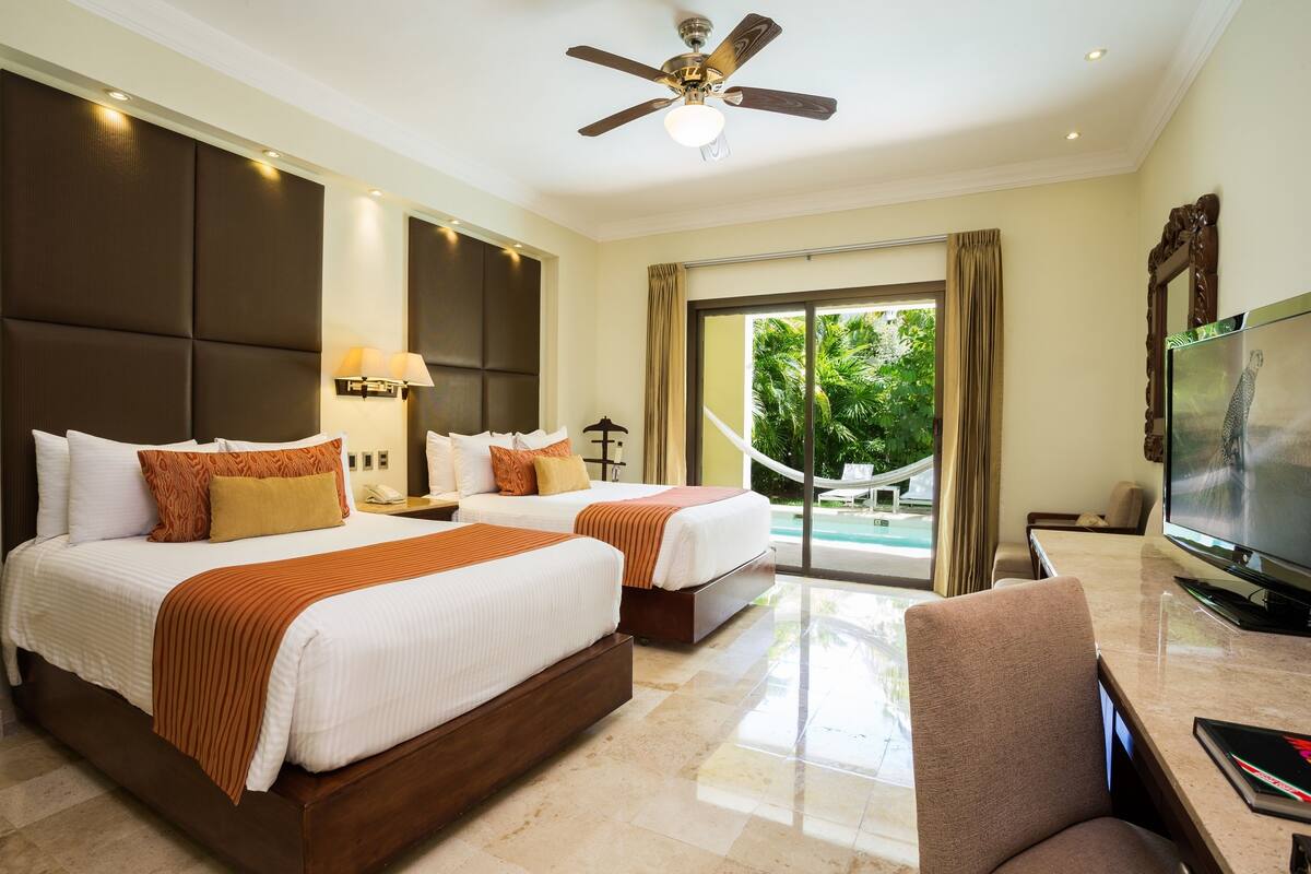 A hotel room with two beds, a TV, a ceiling fan, and a view of the outdoors.