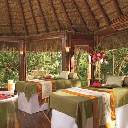 A room with two beds, a bathtub with rose petals, and a view of lush greenery.