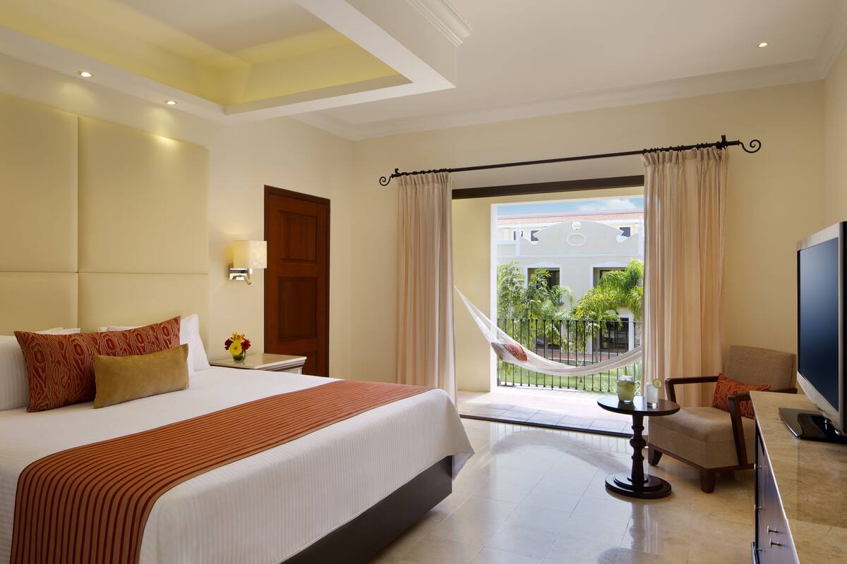 A hotel room with a large bed, a TV, a balcony with a view of the outside, and a small table with a chair.