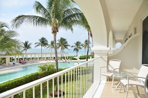 A balcony with white chairs and a pool area with palm trees and lounge chairs.