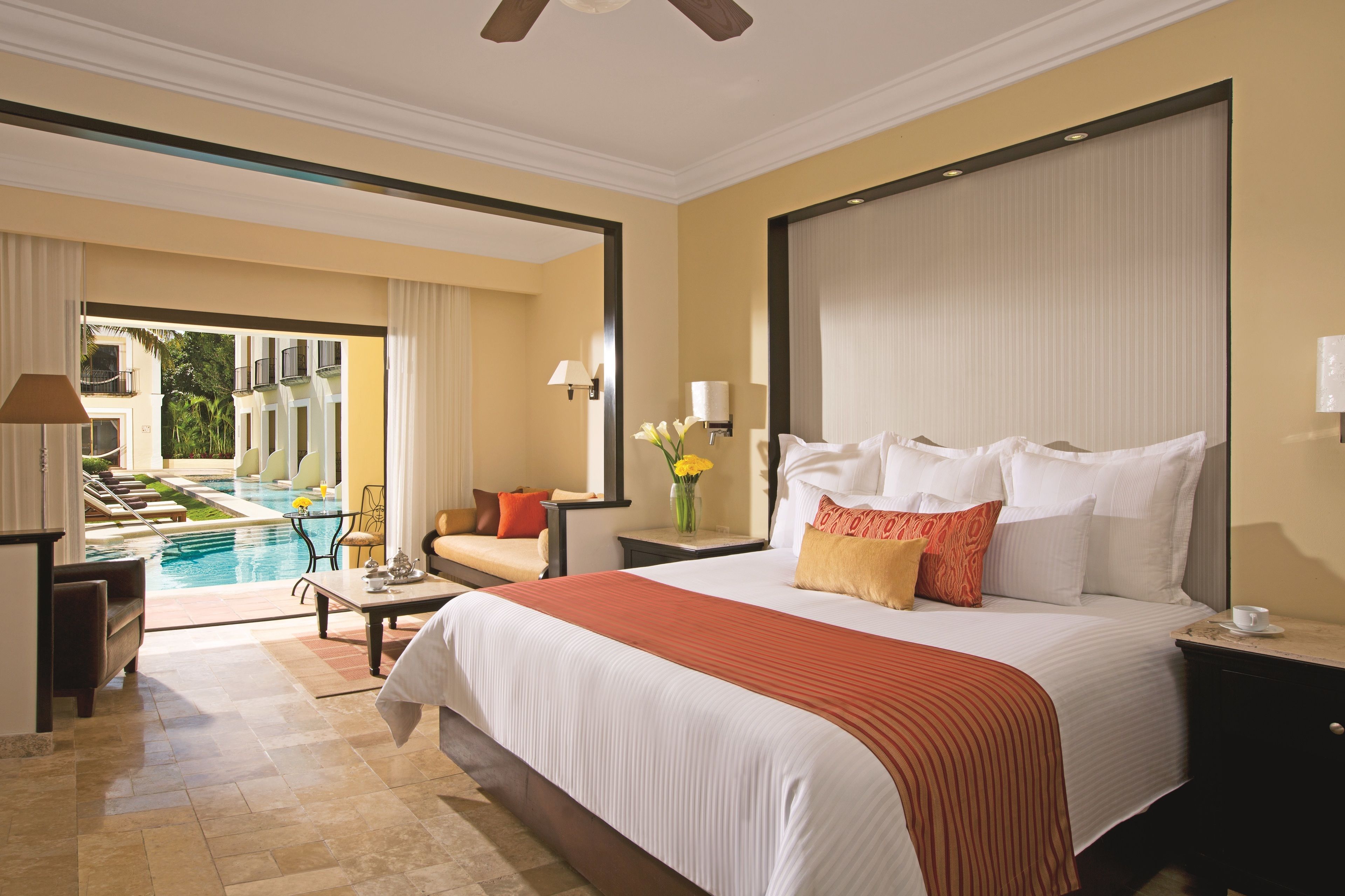 A hotel room with a large bed, a view of a swimming pool, a dining area with a table and chairs, and a separate seating area with a sofa.