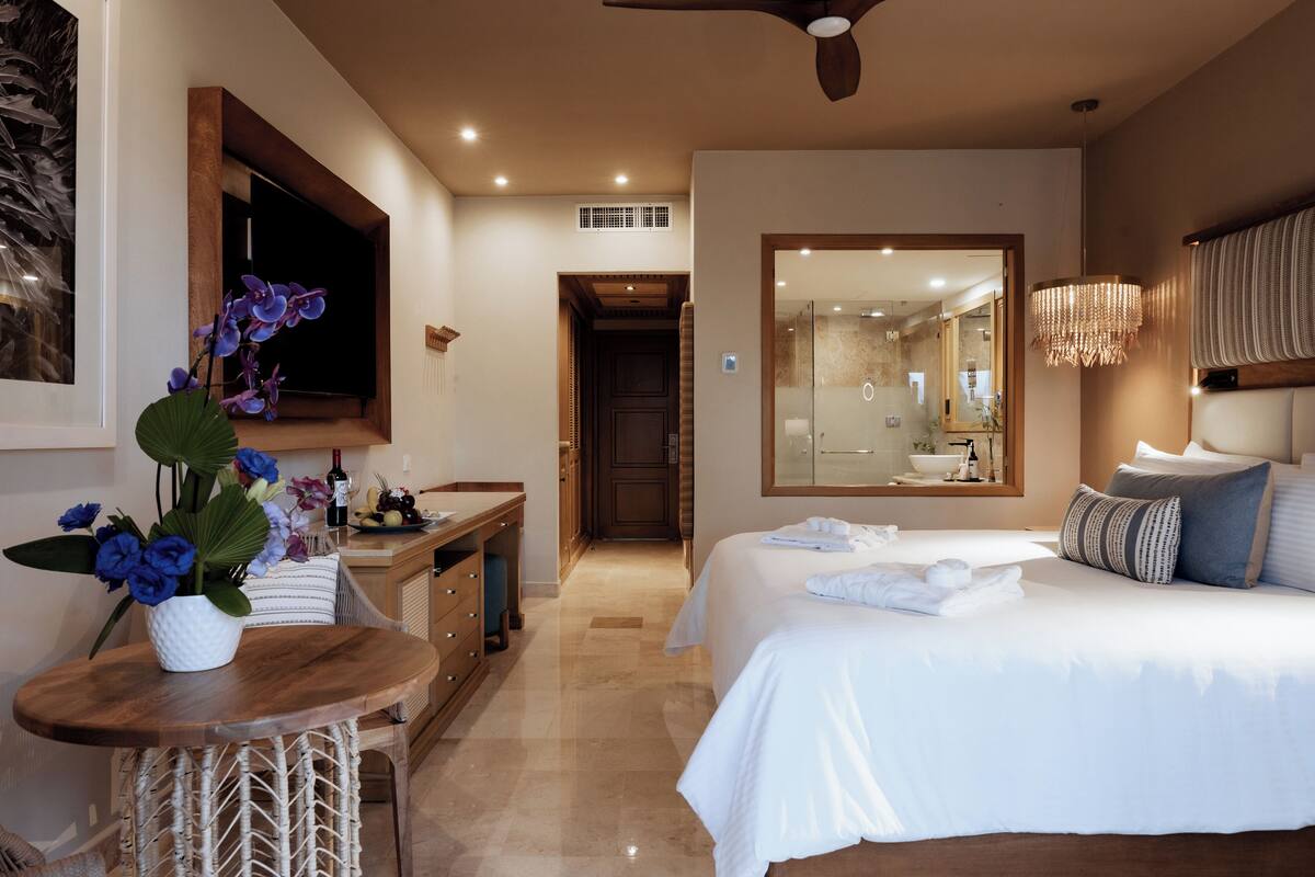 A modern hotel room with a large bed, a desk with a vase of flowers, a television, and a bathroom visible through an open door.