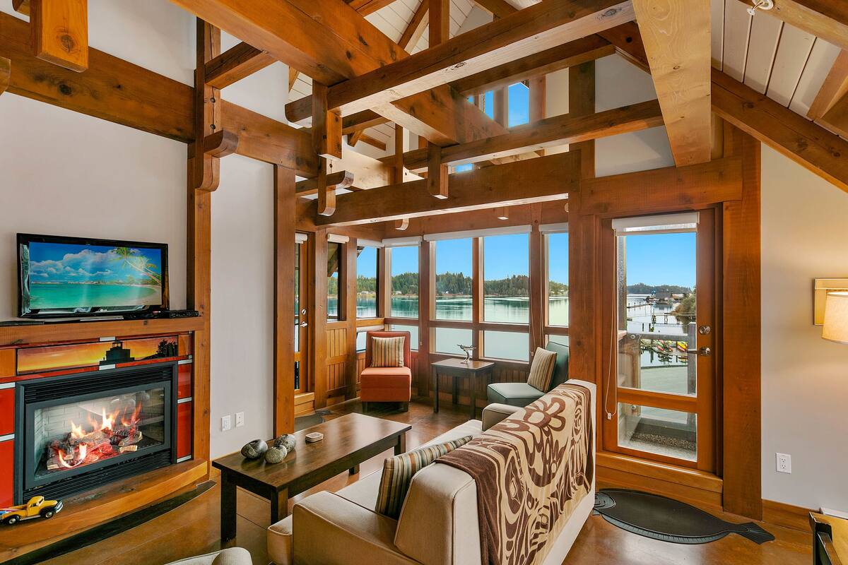 Un salón amplio con chimenea, asientos cómodos, ventanas grandes con vistas a un lago y vigas de madera en el techo.