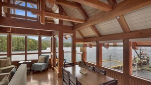 Amplia habitación con techo de madera, ventanas grandes que ofrecen vistas a un lago y un muelle, y un comedor con mesa y sillas.