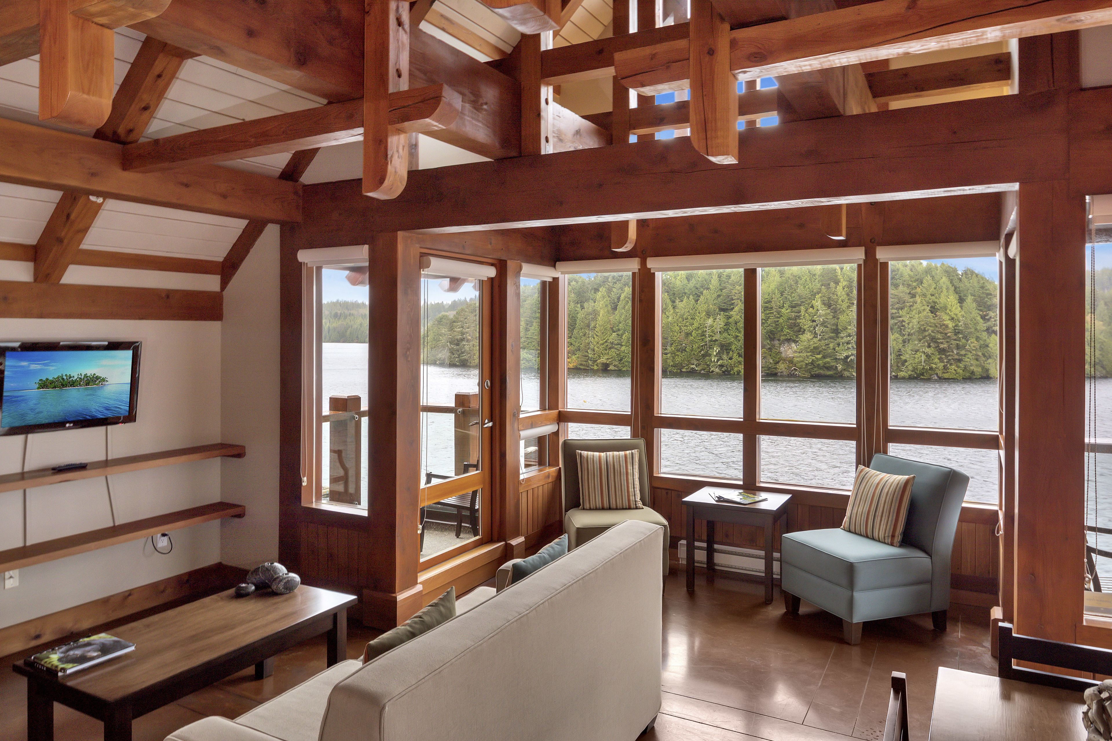 A cabin interior with a large window overlooking a lake, a sofa, a chair, and a television on the wall.