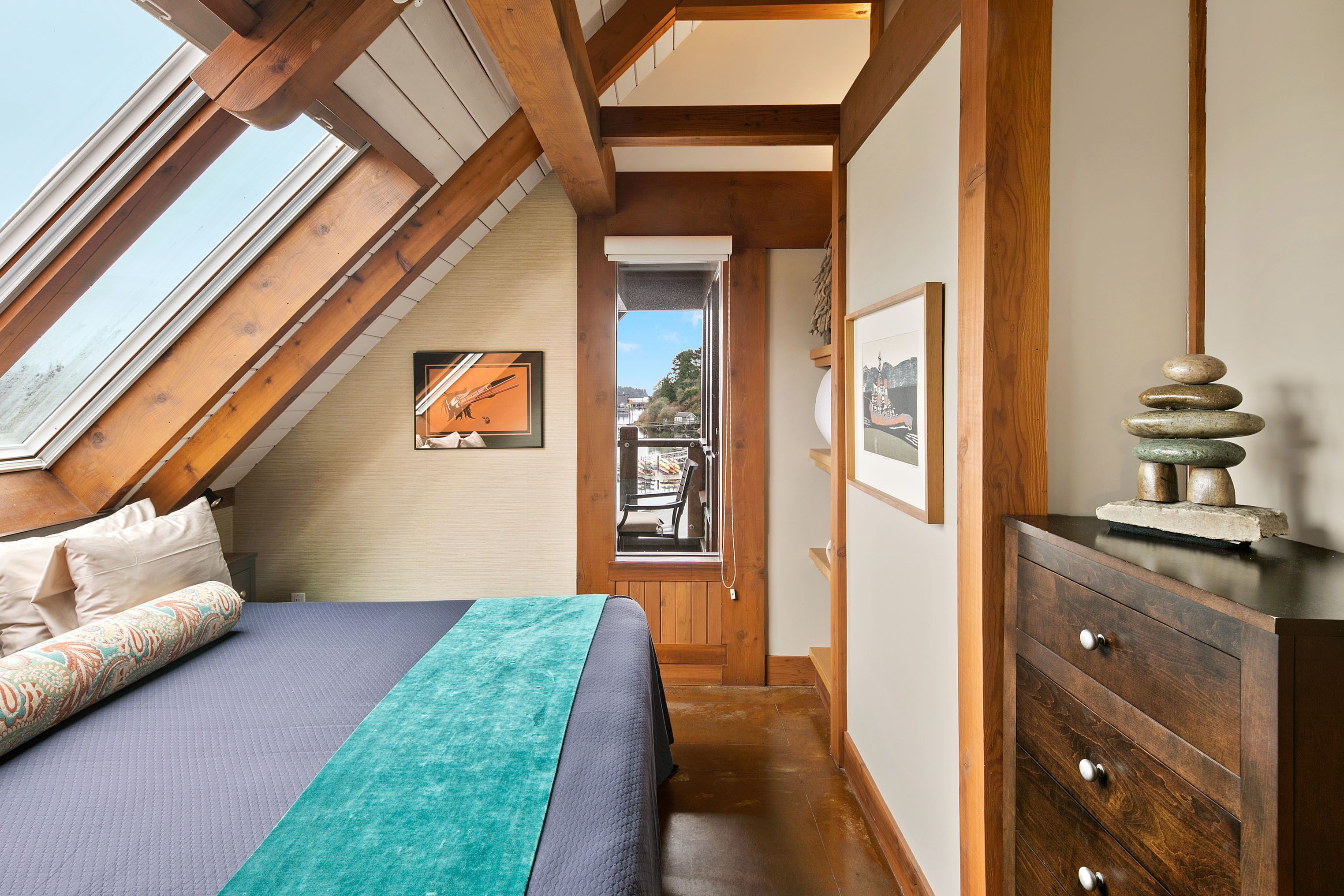 Une chambre avec un plafond en pente, des poutres en bois, un lit avec une literie à motifs, une commode et des œuvres d’art encadrées.
