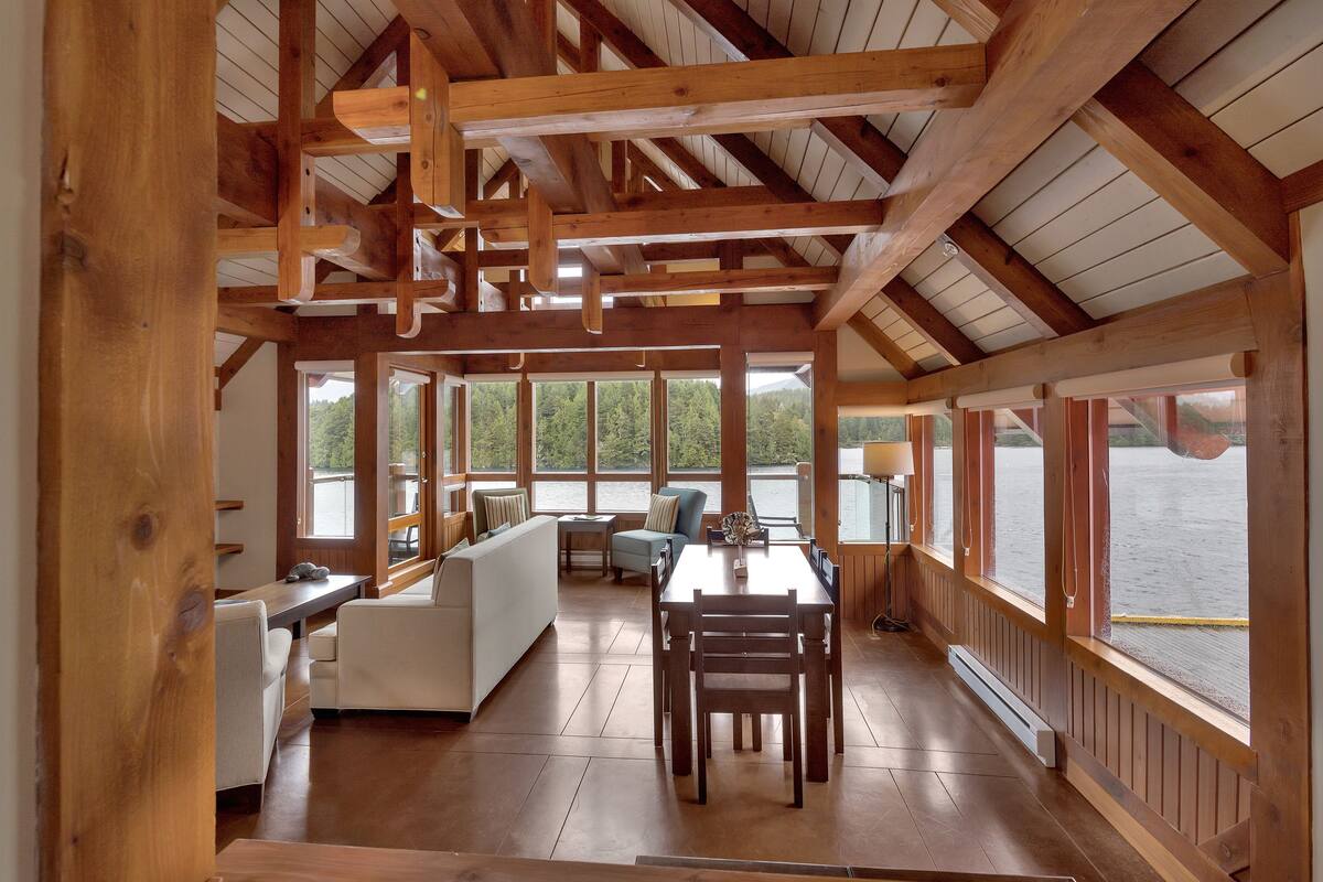 Una habitación amplia con techo de madera, una mesa de comedor con sillas, un sofá y grandes ventanales que ofrecen vistas al exterior.