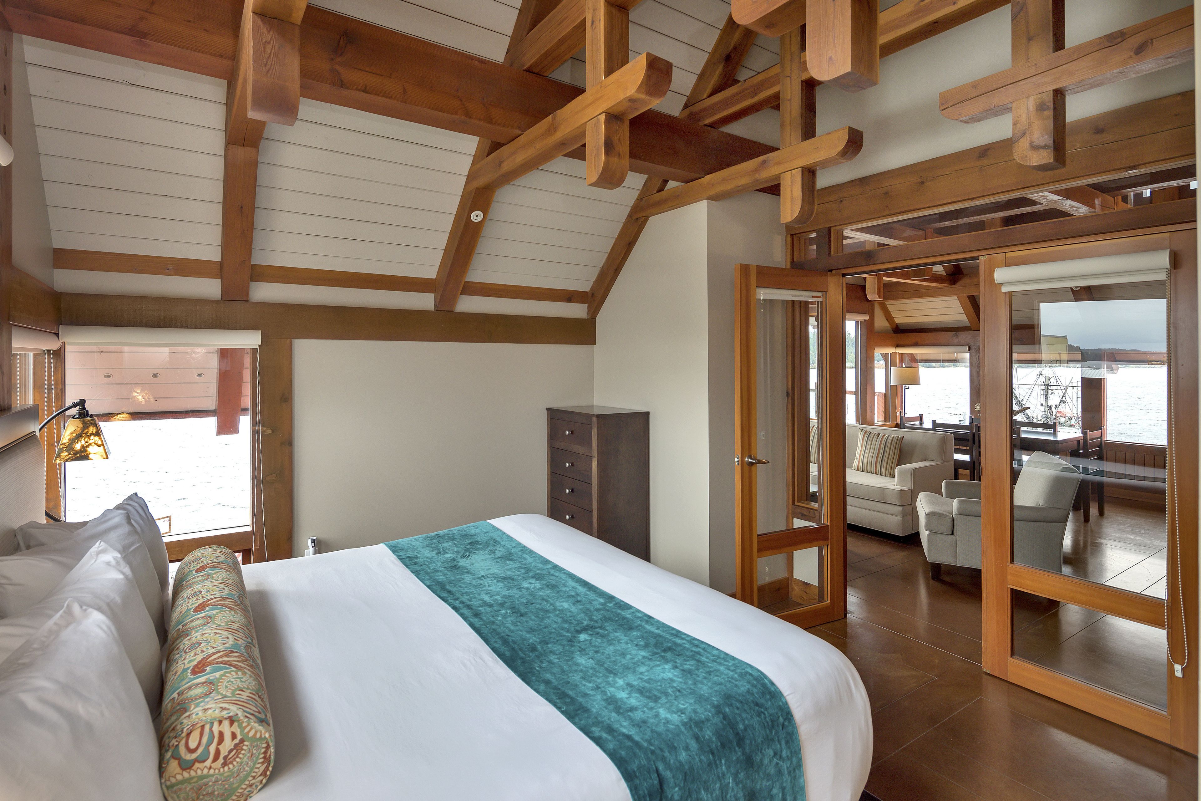 Une chambre spacieuse dotée d’un grand lit, de poutres apparentes en bois au plafond, d’une vue sur l’extérieur et d’un coin salon avec un canapé et des fauteuils.