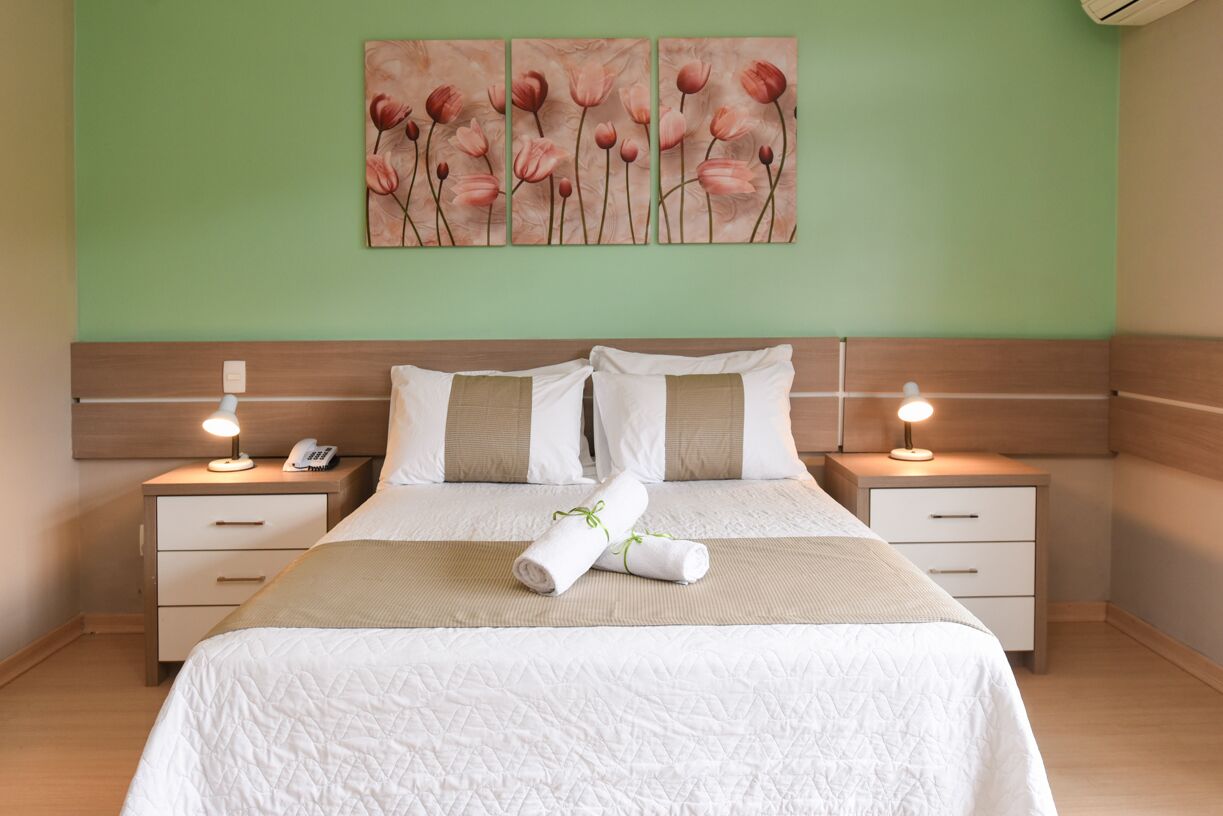 Un lit bien fait, recouvert d’une couette blanche matelassée, avec deux oreillers et une serviette enroulée posée sur une tête de lit en bois. On y trouve deux tables de chevet avec des lampes et un téléphone fixé au mur.