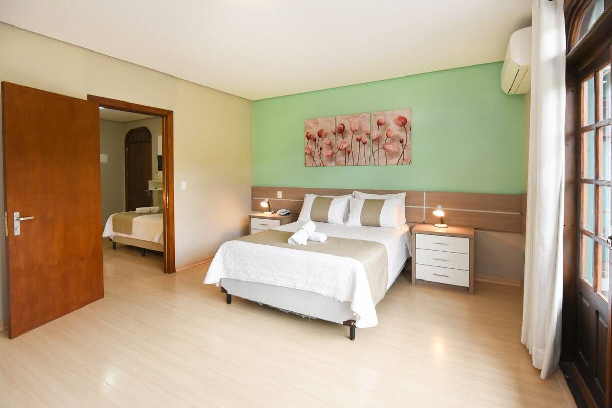 A bedroom with a bed, bedside tables, a nightstand, a wall-mounted air conditioner, and a door leading to another room.