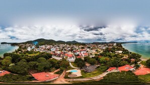 An aerial view of a coastal town with a beach, houses, and a mountainous backdrop. - Pousada Parque Praia (Penha)