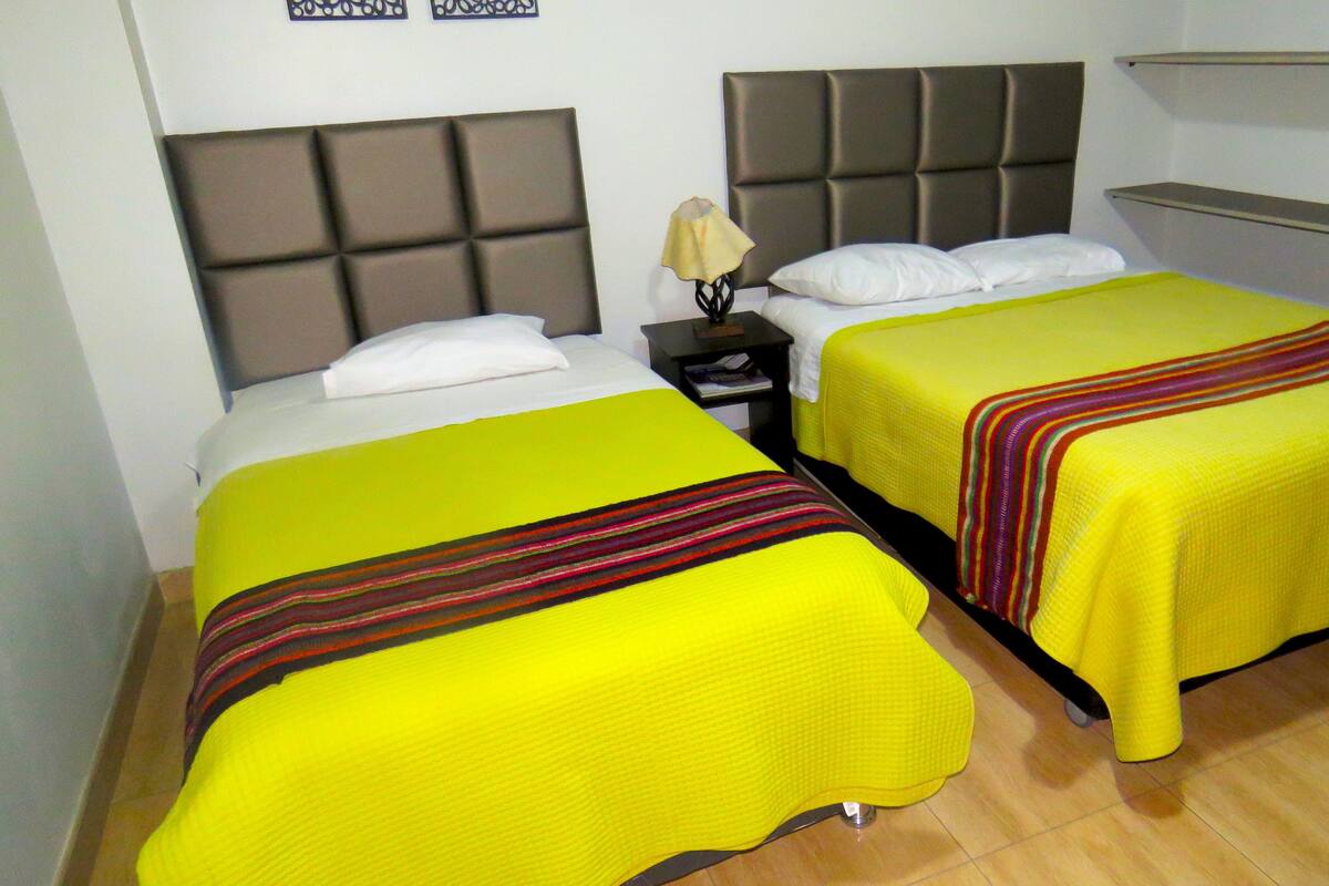 A hotel room with two beds, each with a yellow bedspread and a striped blanket. There is a nightstand with a lamp and a book. The room has a modern design with a grey headboard and a shelf above the beds.