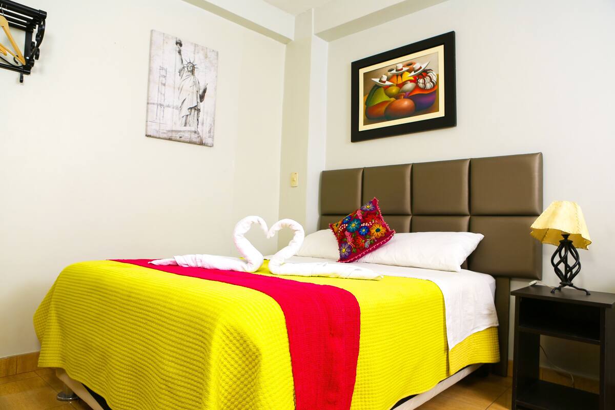 A neatly made bed with a bright yellow and red bedspread, a pillow with a colorful floral pattern, and two white towels arranged in a heart shape.