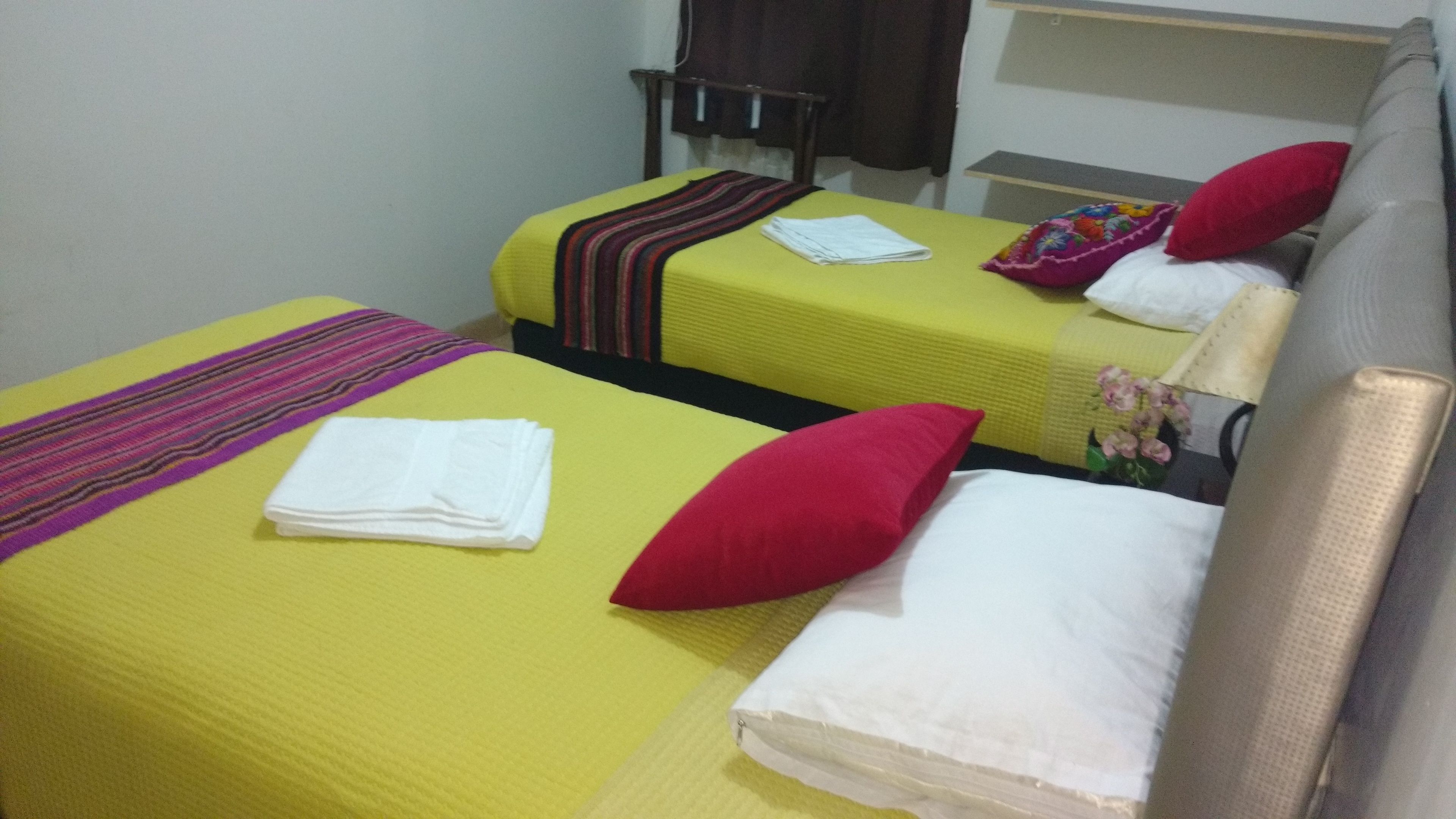 Habitación de hotel con dos camas, cada una con una colcha amarilla y una almohada roja. Una cama tiene una manta a rayas y la otra, una toalla blanca doblada.