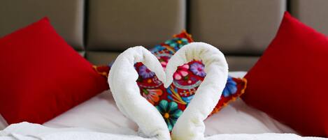 Una cama con toallas blancas dispuestas en forma de corazón, almohadas rojas y una manta floral colorida.
