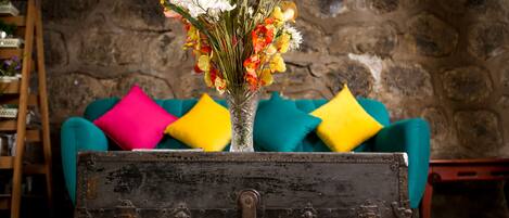 A rustic wooden chest with a glass vase containing a variety of flowers, placed on a tiled floor in front of a teal sofa with colorful cushions.