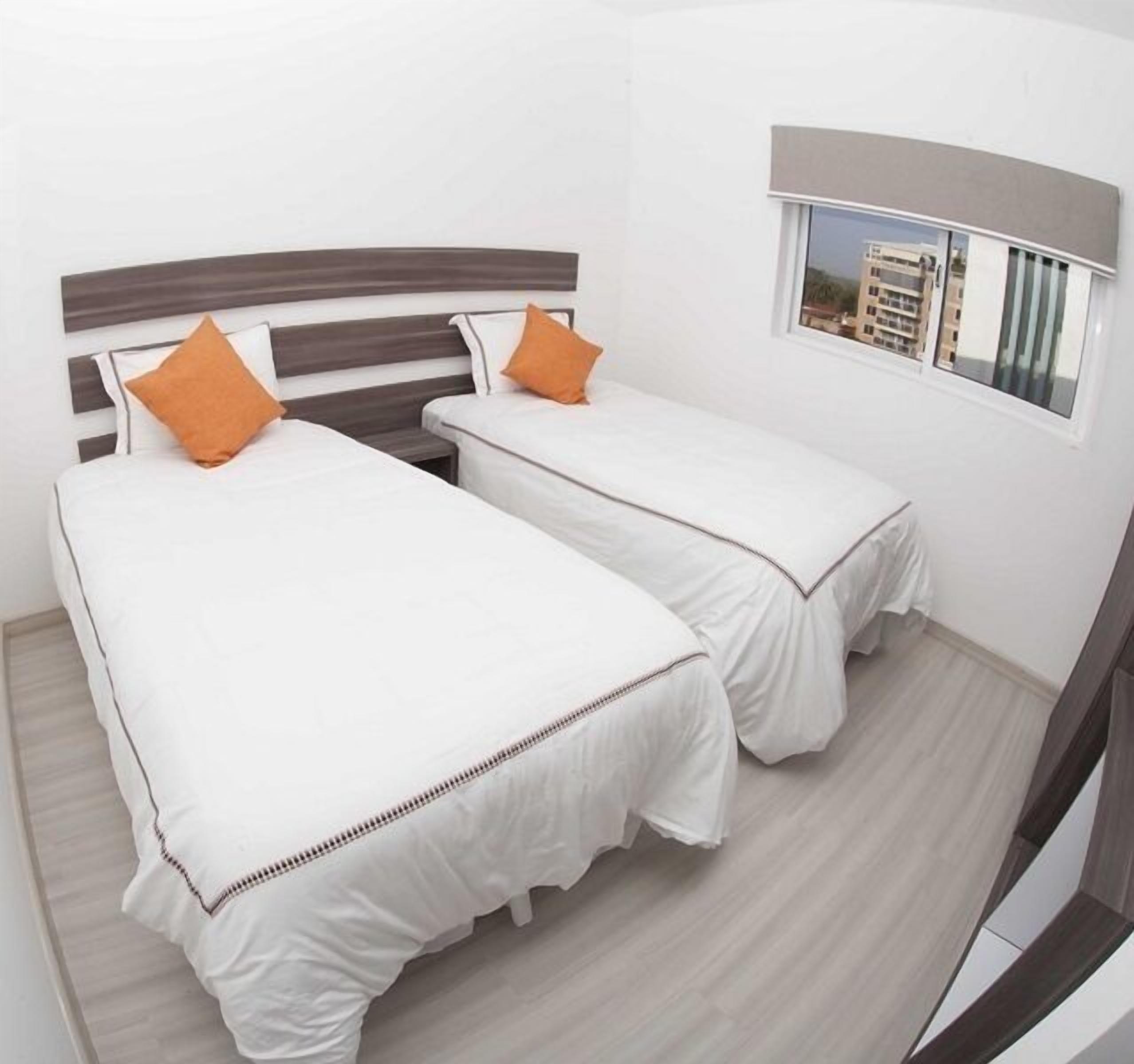 Une chambre d’hôtel avec deux lits simples, chacun recouvert d’une couvre-lit blanche et agrémenté d’un oreiller orange. La chambre est dotée d’un plancher en bois et d’une fenêtre donnant sur des immeubles.