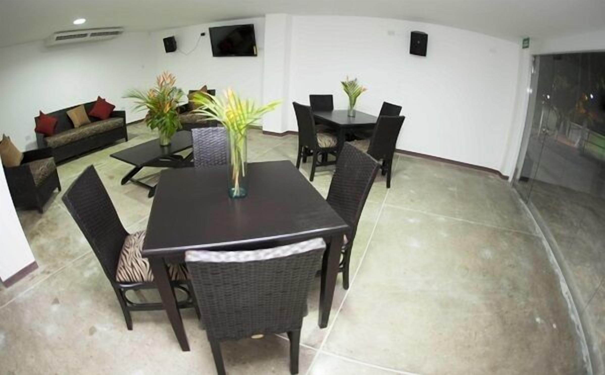 A dining area with a dark wooden table and chairs, surrounded by white walls and a tiled floor. There is a sofa with cushions in the background and a potted plant on the table.