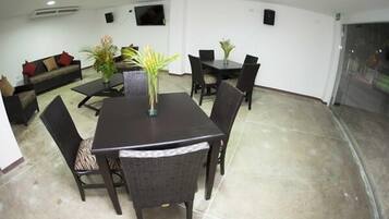A dining area with a dark wooden table and chairs, surrounded by white walls and a tiled floor. There is a sofa with cushions in the background and a potted plant on the table.