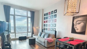 A modern living room with a sofa, a dining table with chairs, a TV, and a large window with curtains. - Monki-Di Executive Suites - Alura (Calgary)